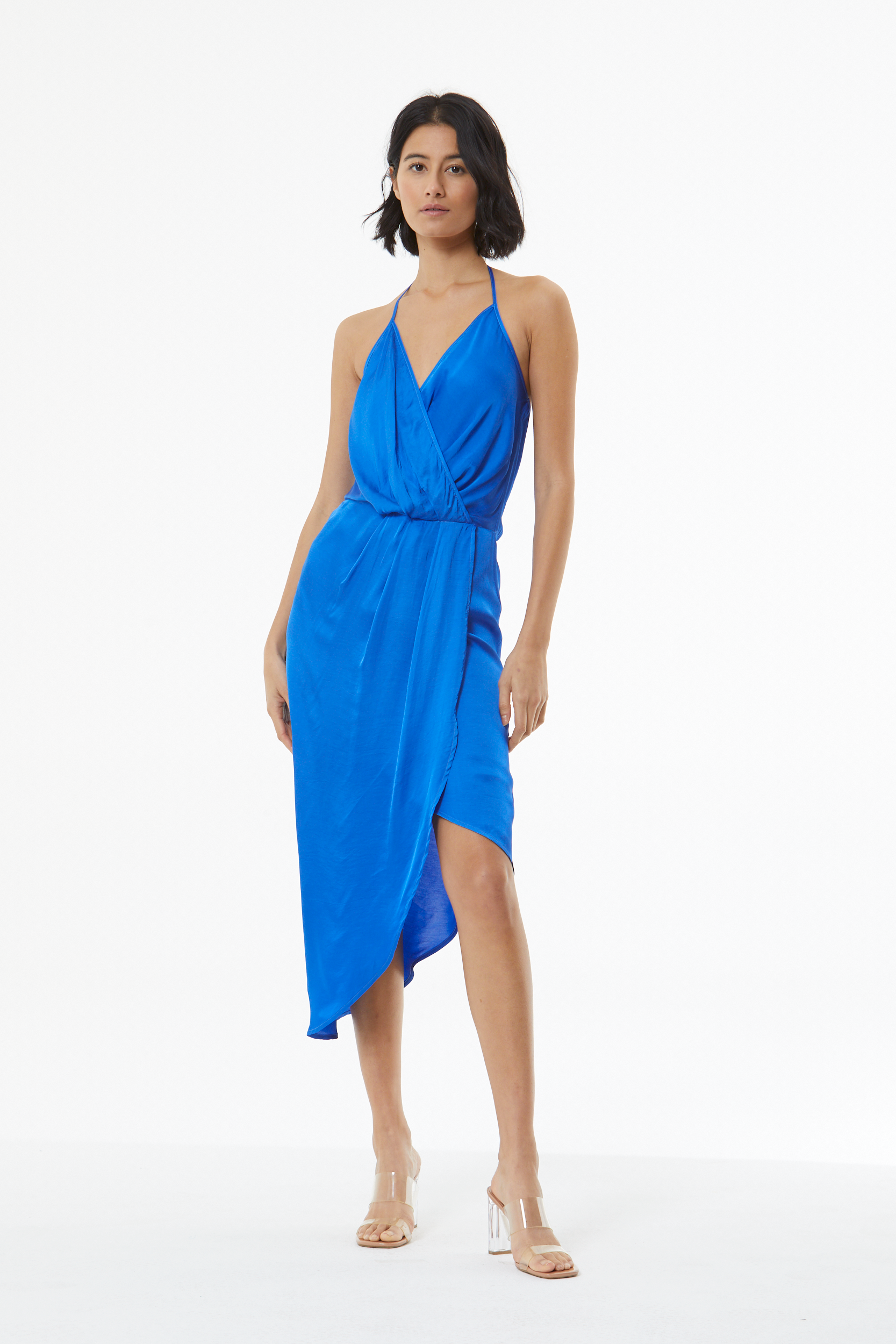 A woman modeling a bright royal blue wrap-style midi dress with thin straps, a draped neckline, and a gathered waist, photographed against a clean white studio background.