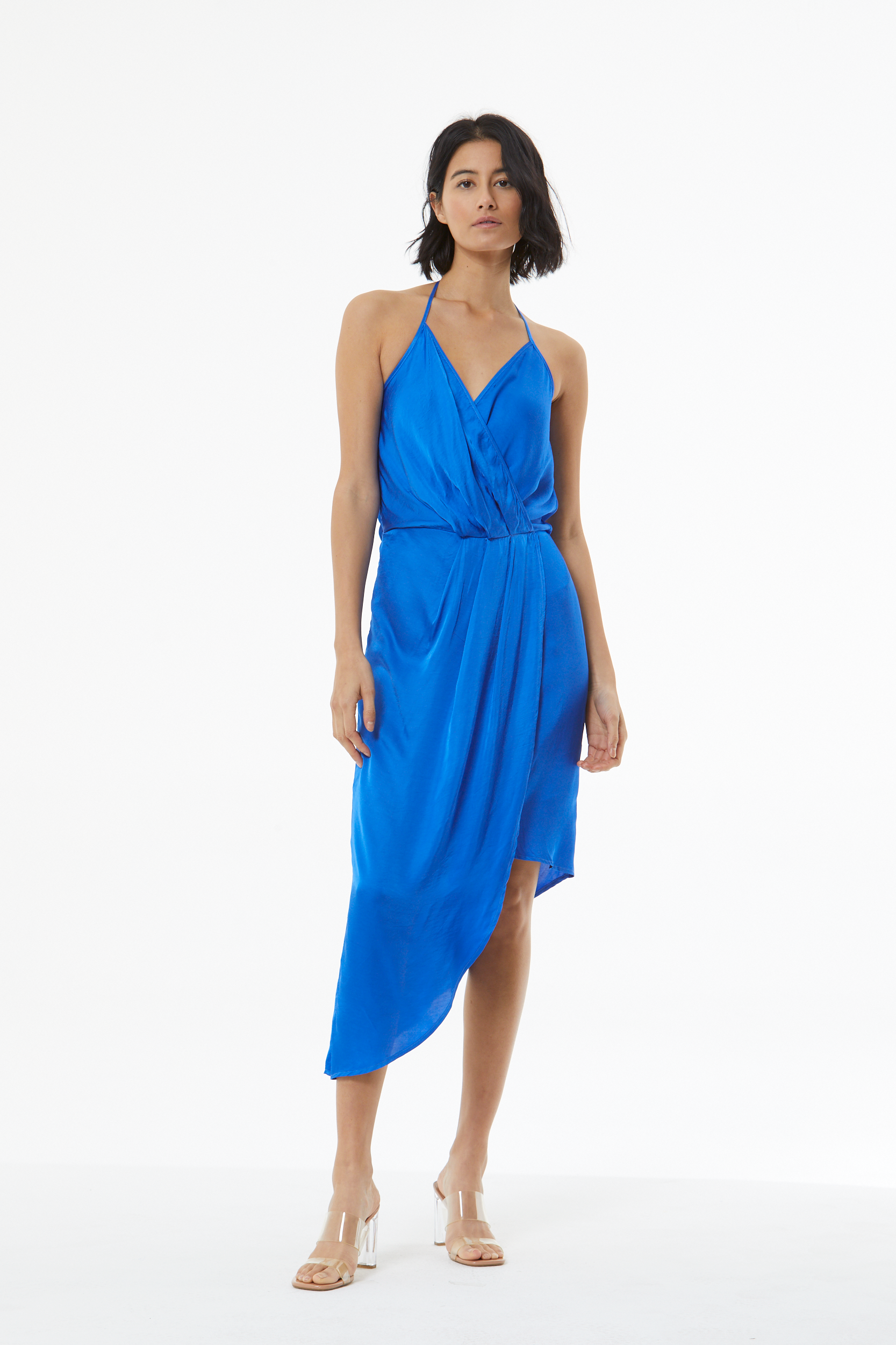 A woman modeling a bright royal blue halter midi dress with a draped, wrap-style bodice and asymmetrical hem, paired with clear high-heel sandals, against a plain white studio background.