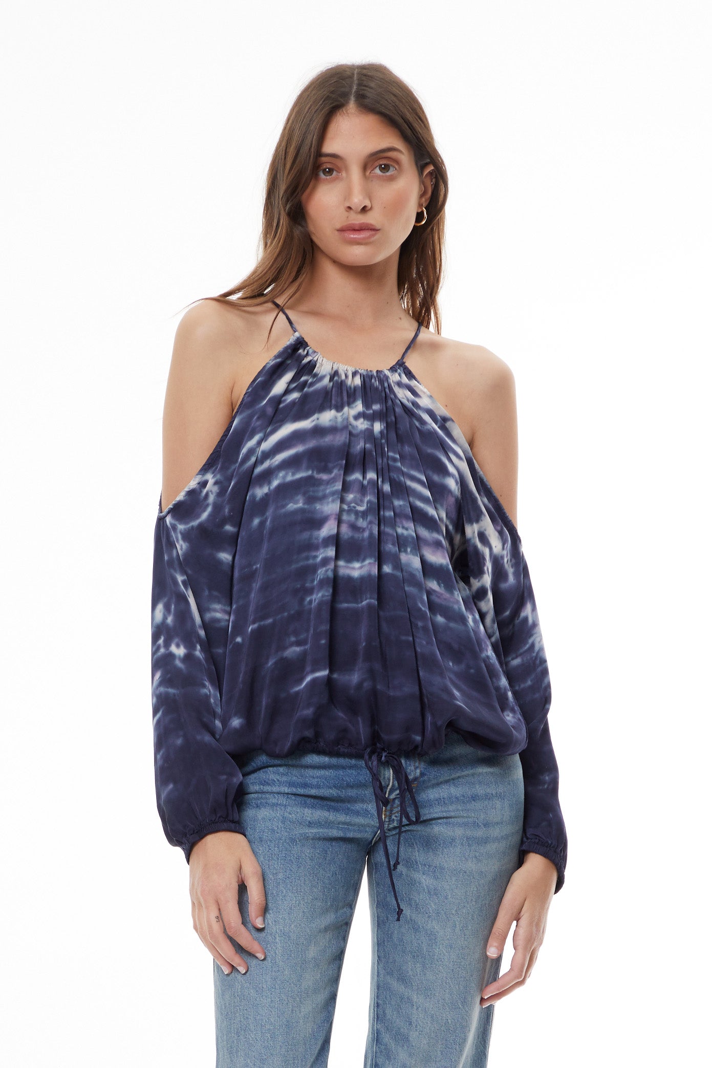A woman modeling a dark blue and white tie-dye cold-shoulder top with long balloon sleeves, paired with light-wash blue jeans and black ankle boots, against a plain white studio background.