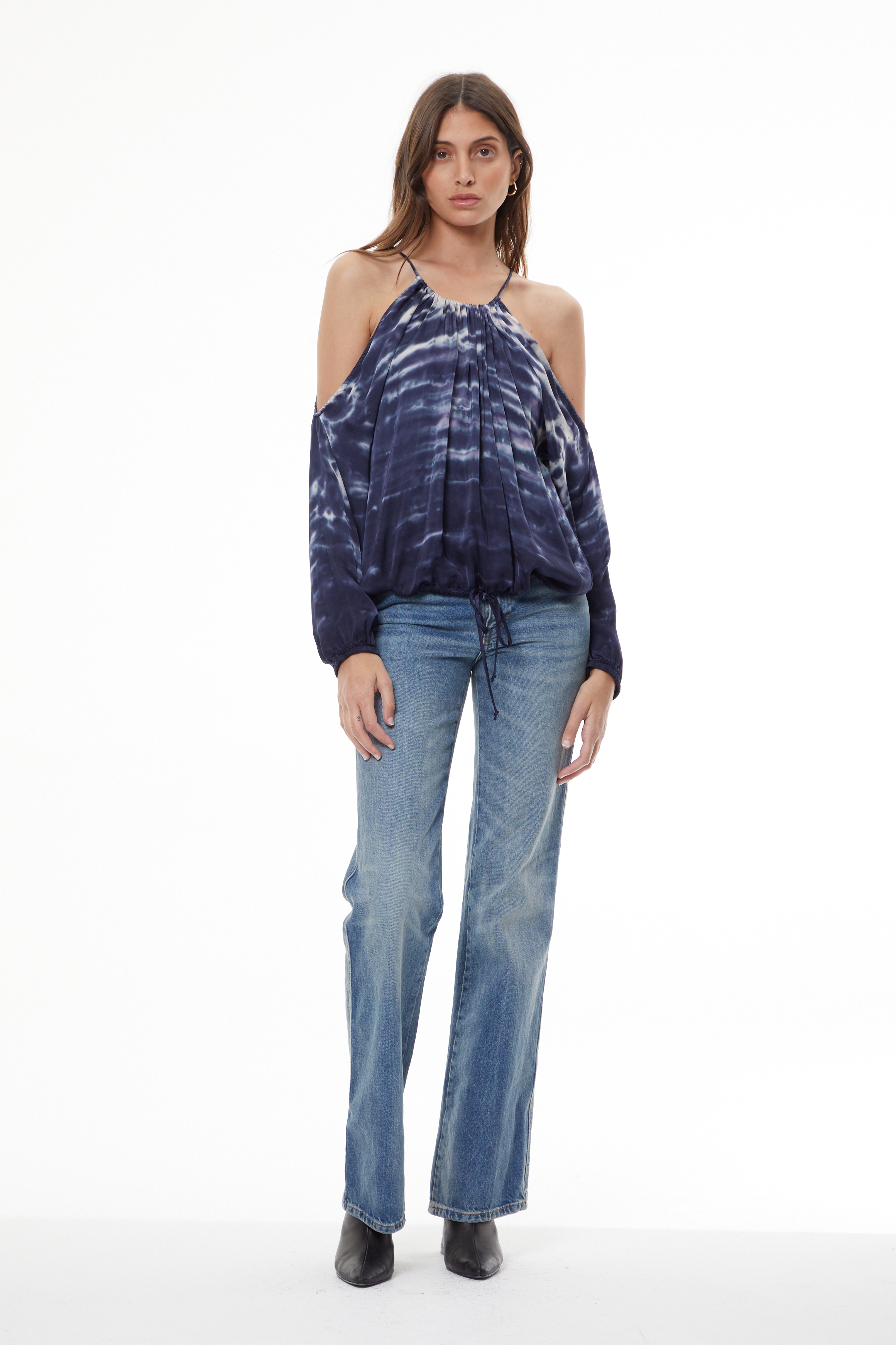 A woman modeling a dark blue and white tie-dye cold-shoulder top with long balloon sleeves, paired with light-wash blue jeans and black ankle boots, against a plain white studio background.