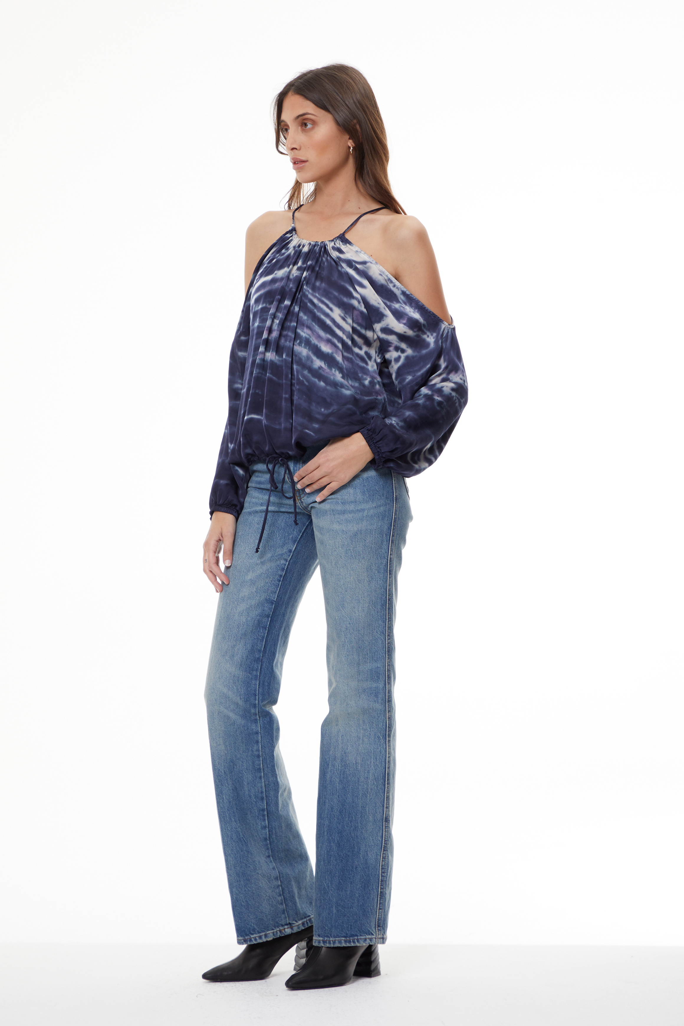 A woman modeling a dark blue and white tie-dye cold-shoulder top with long balloon sleeves, paired with light-wash blue jeans and black ankle boots, against a plain white studio background.