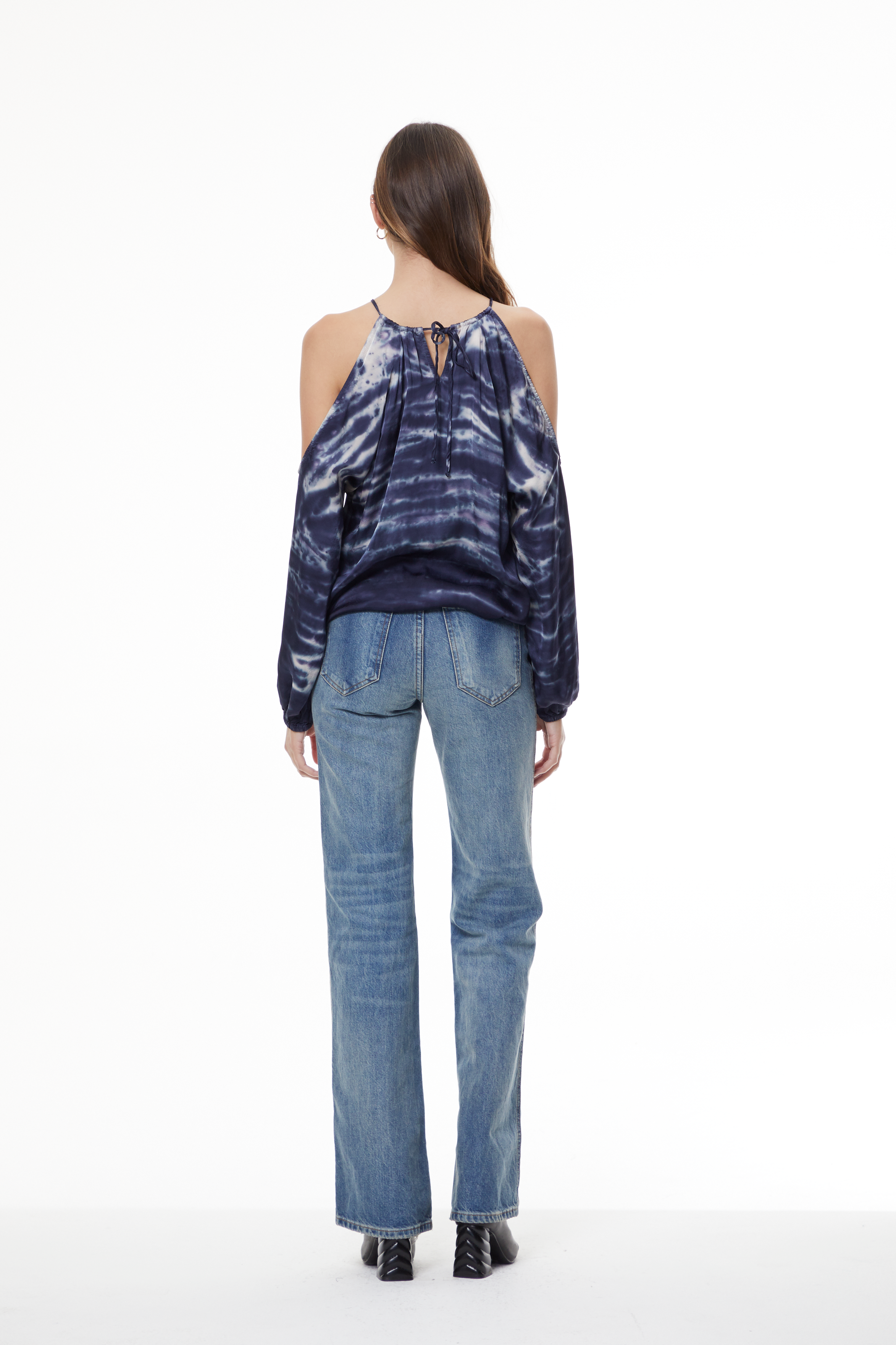 A woman viewed from the back wearing a dark blue and navy tie-dye cold-shoulder top with blue jeans and black shoes, against a plain white studio background.
