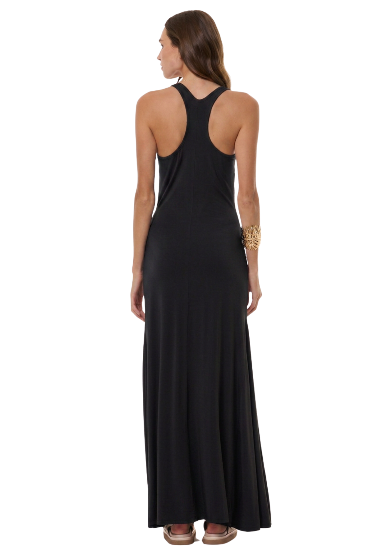 A woman viewed from the back wearing a sleeveless black maxi dress with a wide shoulder cut and a deep open back, paired with brown sandals and a gold bracelet, against a plain white background.