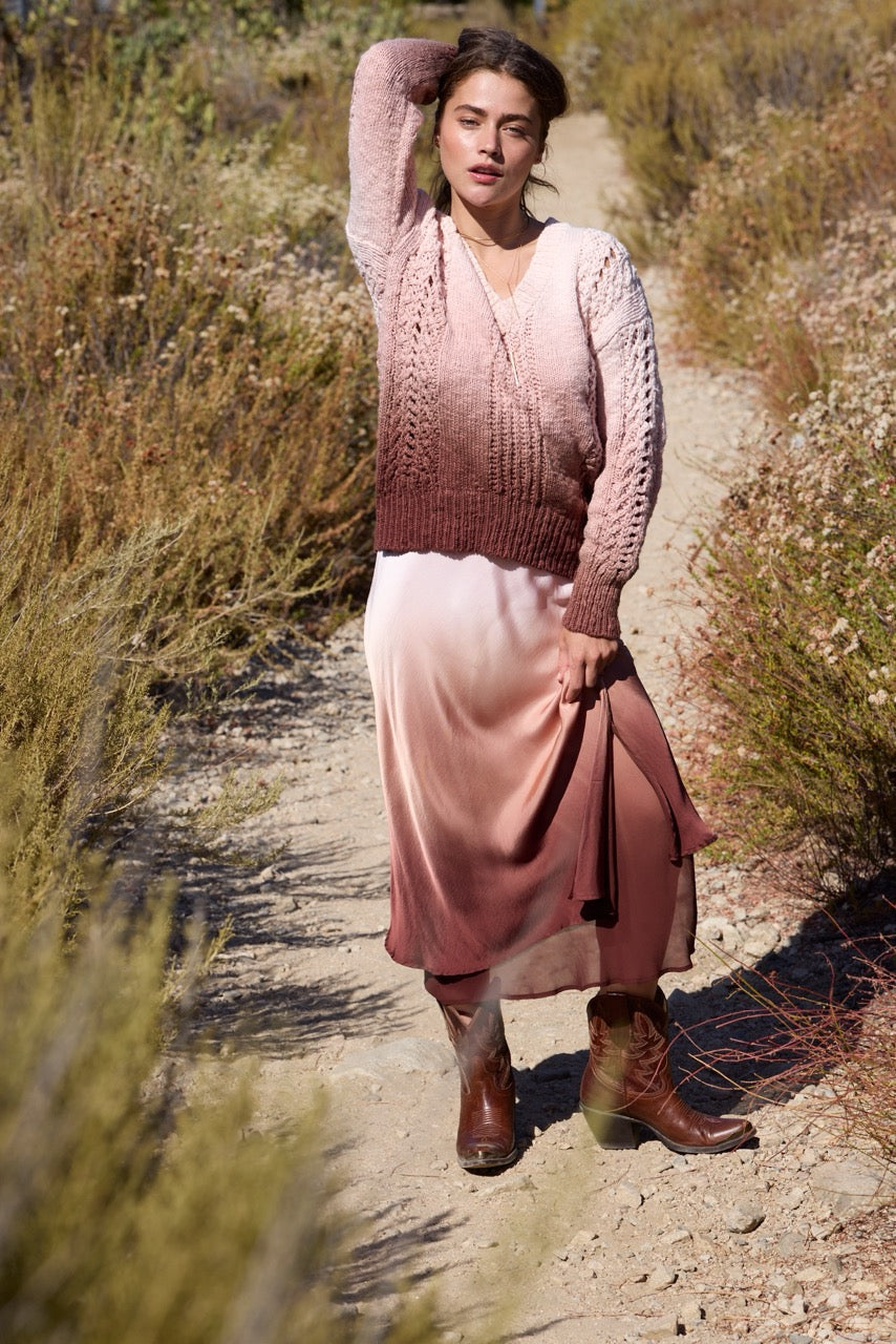 A woman stands on a dirt path outdoors wearing a pink gradient cable-knit sweater vest over a matching pink gradient satin midi skirt, paired with brown leather boots.