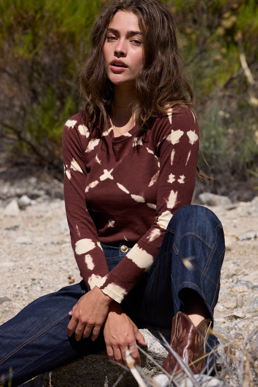 A woman sitting outdoors on a rocky surface, wearing a long-sleeve brown and cream tie-dye knit top, blue jeans, and brown boots.