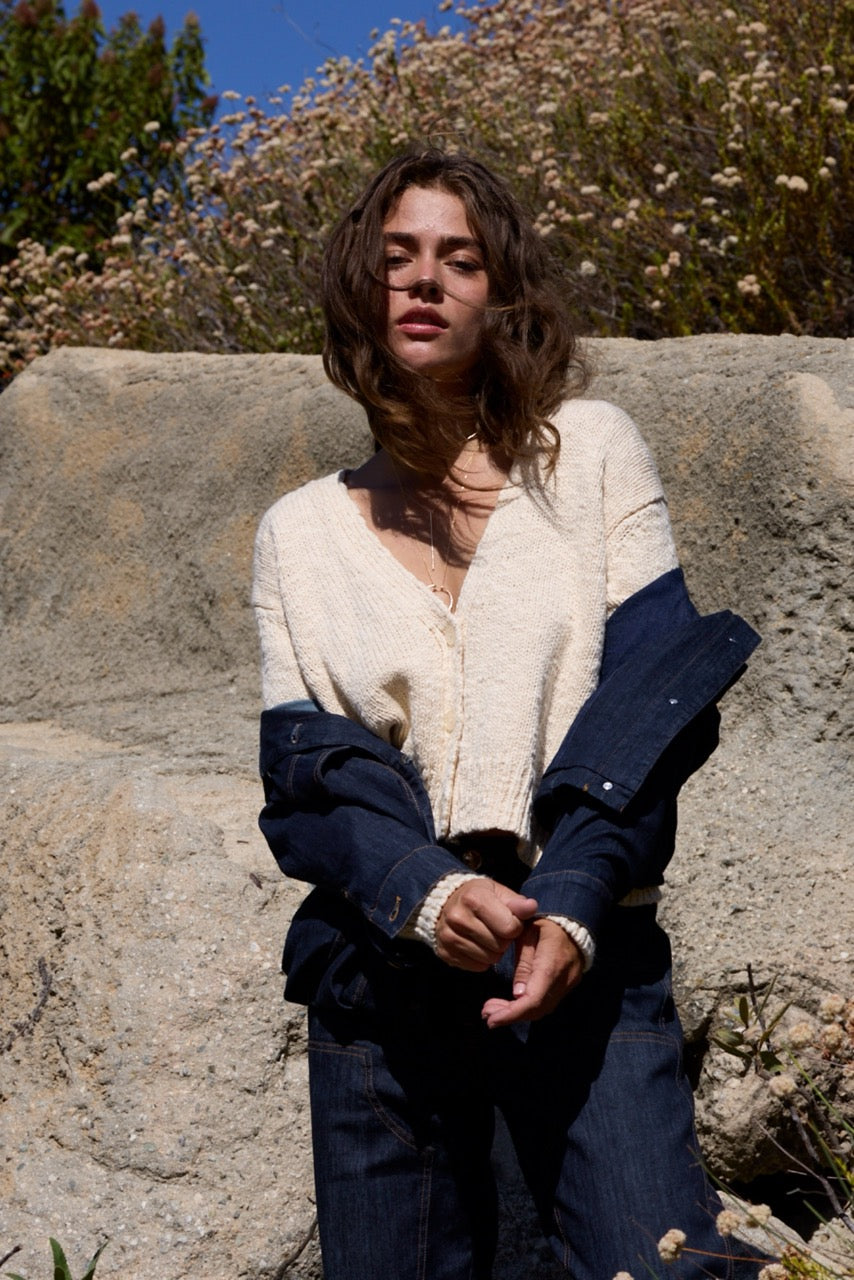 A woman stands outdoors against a large rock with wildflowers and trees in the background, wearing a cream knit cardigan layered over a dark navy denim jacket and dark blue jeans.