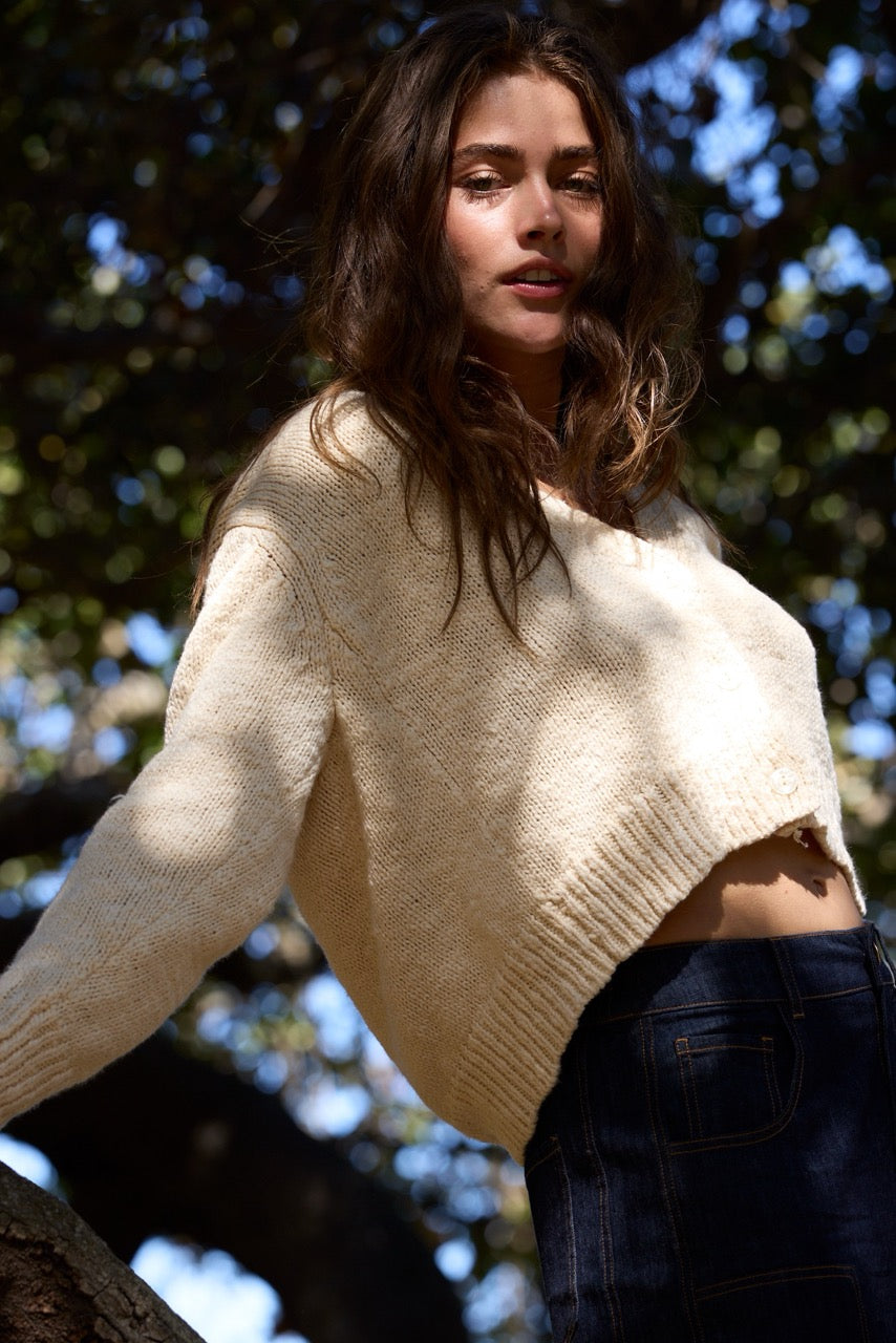 A woman outdoors wearing a cream-colored knit cropped sweater and dark blue jeans, with dappled sunlight filtering through trees.