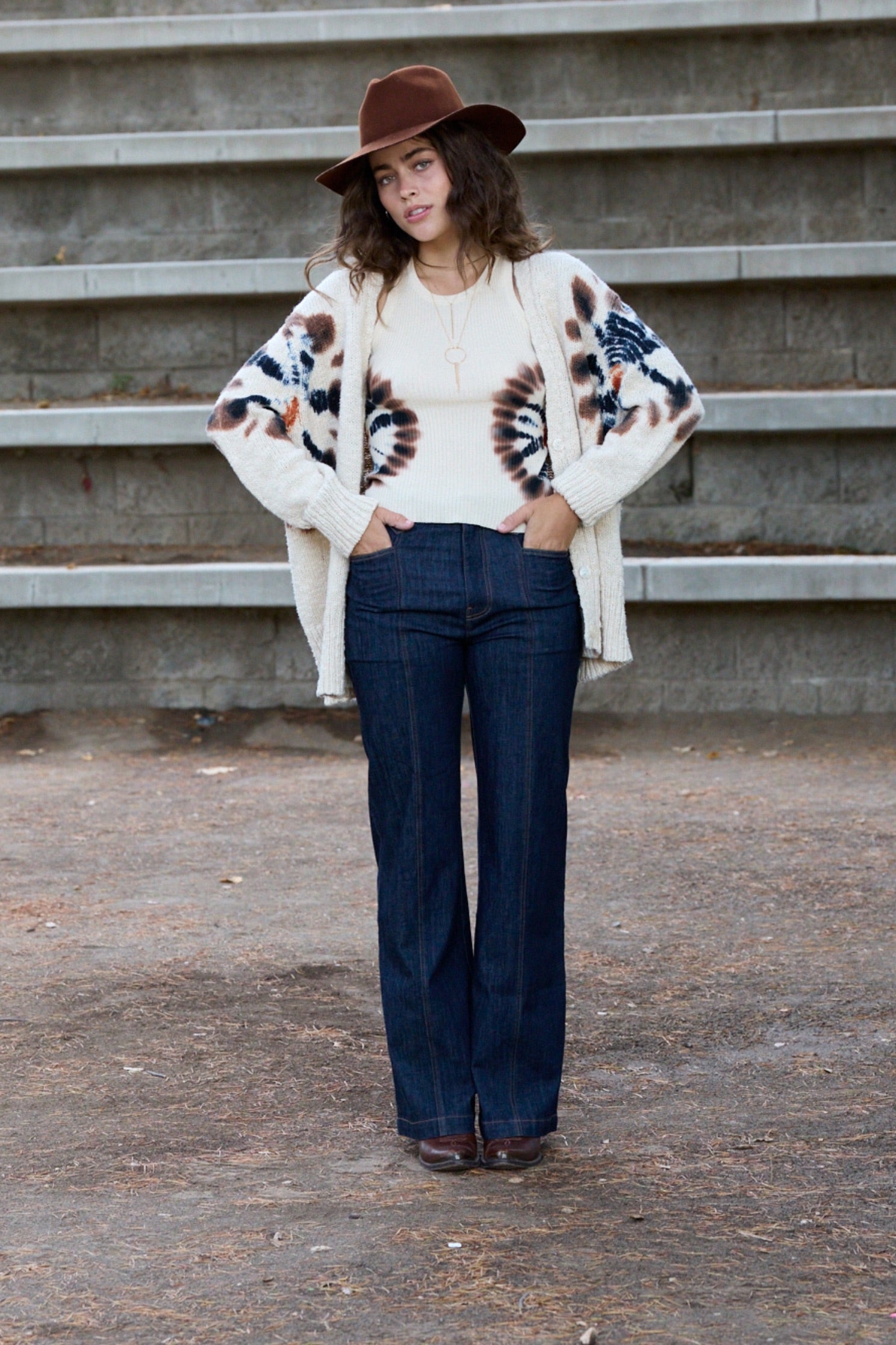 A woman stands outdoors in front of concrete steps wearing a brown wide-brim hat, a cream knit cardigan with brown and blue abstract patterns, a cream ribbed top, dark blue flared jeans, and brown shoes.