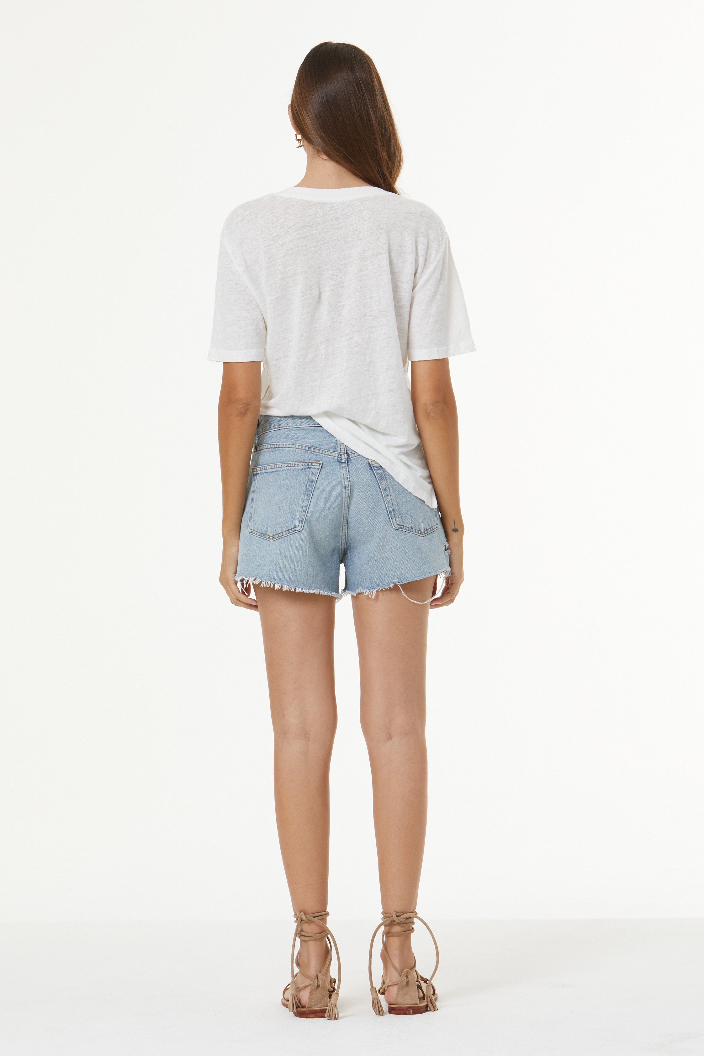 A woman shown from the back wearing a light gray oversized t-shirt, light-wash denim cutoff shorts, and brown lace-up heeled sandals against a plain white studio background.