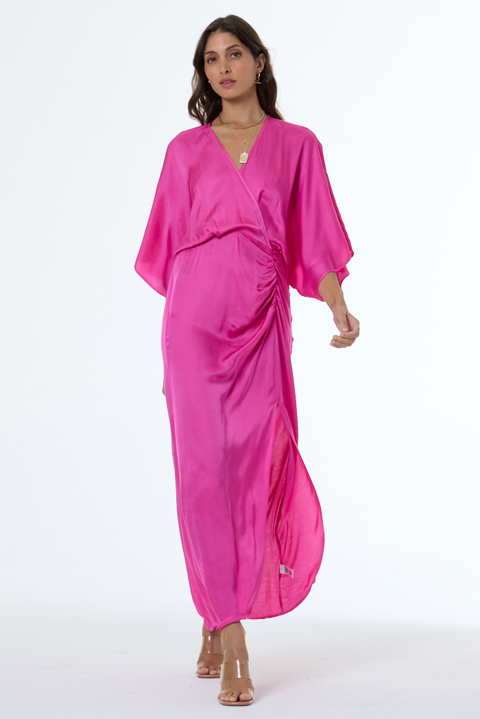 A woman modeling a bright magenta pink satin kaftan dress with wide sleeves and a gathered waist, set against a plain white studio background.