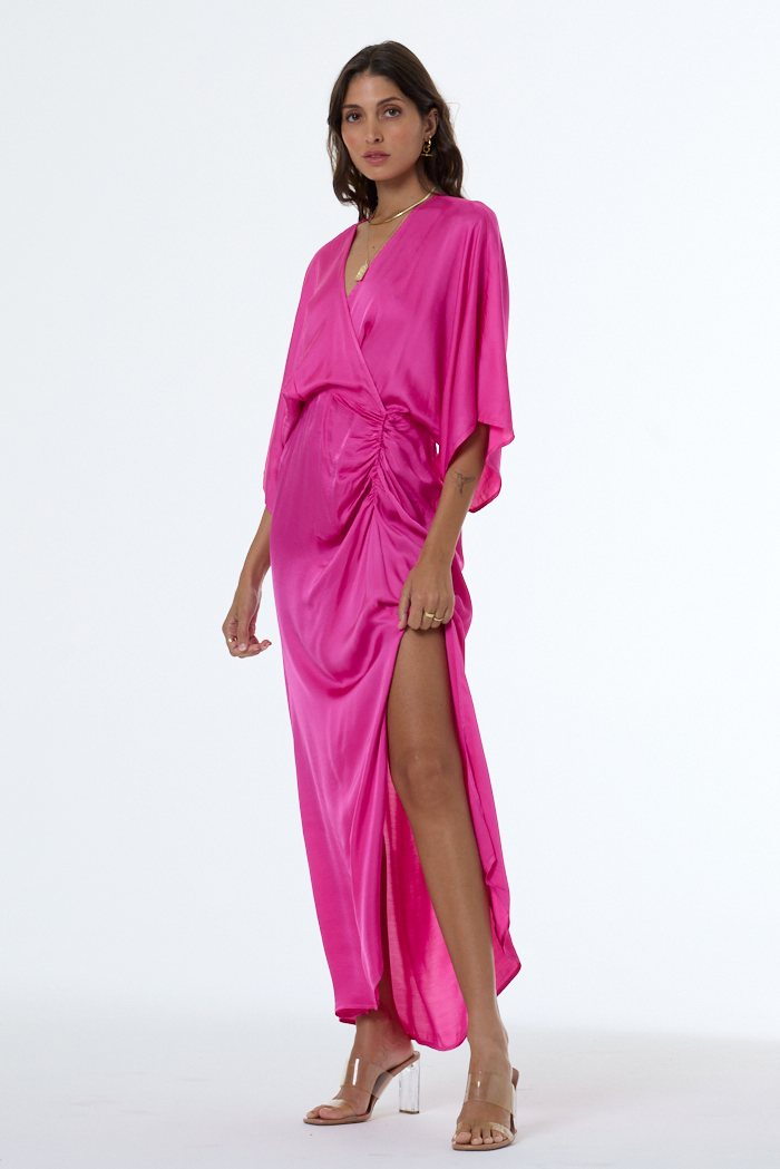 A woman modeling a bright magenta satin wrap dress with a high thigh slit, draped waist, and wide draped sleeves, paired with clear high-heeled sandals against a white studio background.