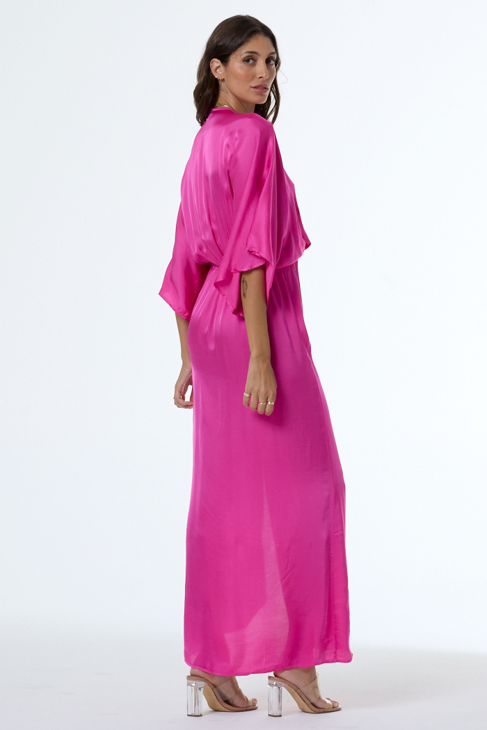 A woman modeling a bright magenta pink maxi dress with three-quarter flutter sleeves and a self-tie waist, shown from a three-quarters back view against a plain white studio background.