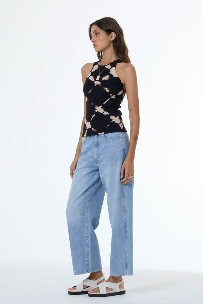 Woman wearing a black sleeveless top with a tie dye pattern and light blue jeans on a white background