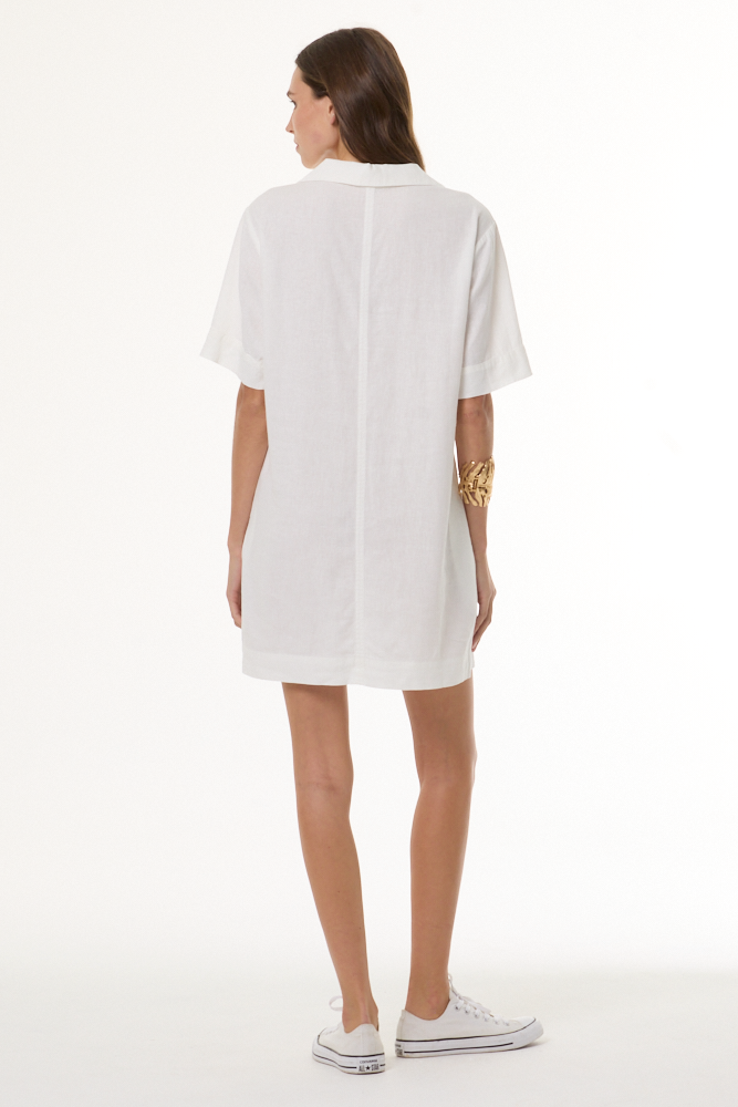 A woman viewed from the back wearing a white short-sleeve button-up shirt dress, paired with white sneakers and a gold bracelet, against a plain white studio background.