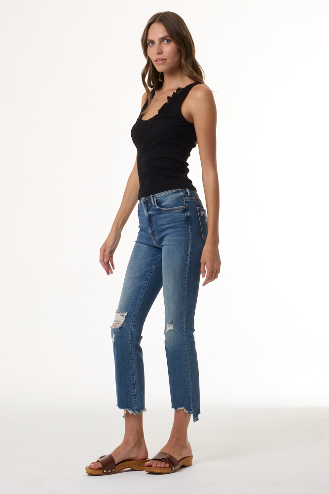 A woman modeling a black sleeveless top and blue cropped denim jeans with distressed details and raw hems, paired with brown slide sandals, against a plain white studio background.