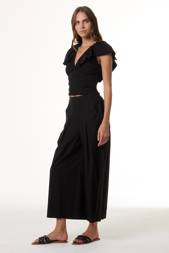 A woman modeling a black two-piece outfit consisting of a ruffled short-sleeve crop top and matching high-waisted wide-leg pants, paired with black slide sandals, against a plain white studio background.