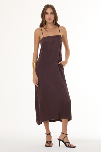 A woman modeling a dark brown midi-length slip dress with thin spaghetti straps, side pockets, and a relaxed drape, paired with black strappy high-heeled sandals against a plain white studio background.