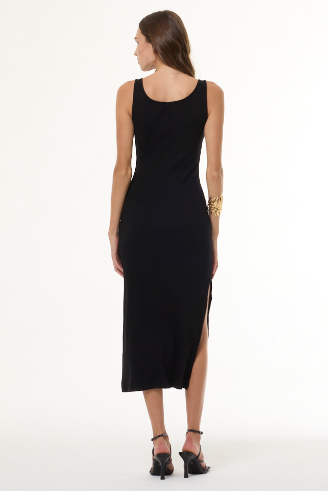 A woman modeling a sleeveless black midi dress with a deep V-neckline, scalloped lace-up detail at the neckline, and a thigh-high side slit, paired with black strappy heels against a plain white studio background.