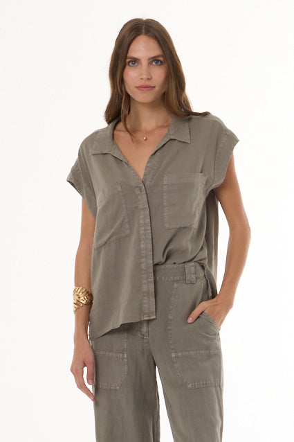 A woman modeling a matching two-piece set consisting of a short-sleeve button-up shirt and coordinating pants in a muted olive-gray color, shot against a plain white studio background.