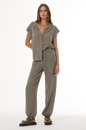 A woman modeling a matching two-piece linen set in a muted olive-gray color, featuring a short-sleeve button-up shirt and wide-leg trousers, paired with dark brown sandals against a plain white studio background.