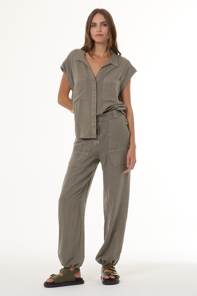 A woman modeling a matching two-piece linen set in a muted olive-gray color, featuring a short-sleeve button-up shirt and wide-leg trousers, paired with dark brown sandals against a plain white studio background.