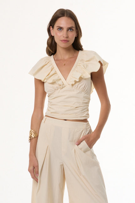 A woman modeling a cream-colored ruffled wrap top with short flutter sleeves and matching high-waisted wide-leg trousers against a plain white studio background.