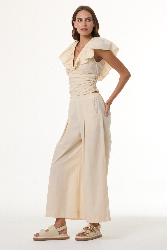 A woman modeling a cream-colored two-piece outfit consisting of a ruffled short-sleeve top and matching wide-leg pants, paired with beige sandals, against a plain white studio background.