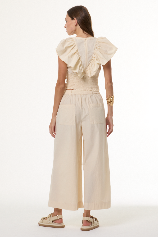 A woman shown from the back wearing a cream-colored two-piece outfit consisting of a ruffled short-sleeve top and matching wide-leg cropped pants, paired with beige sandals against a plain white studio background.