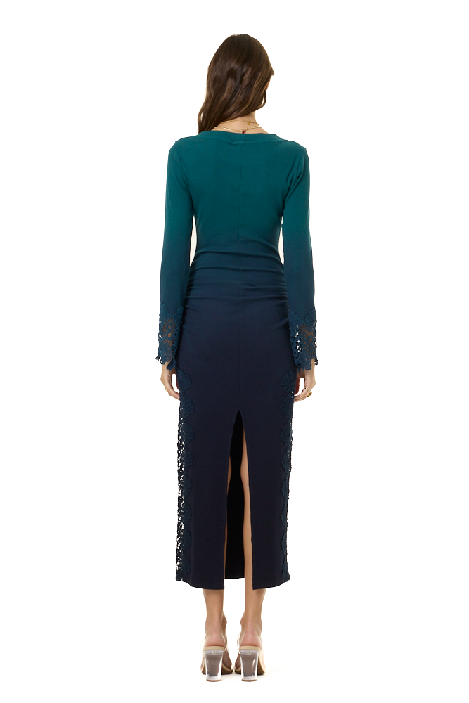 A woman viewed from the back wearing a dark teal long-sleeve top and a navy midi skirt with a front slit and lace trim, standing against a plain white background.