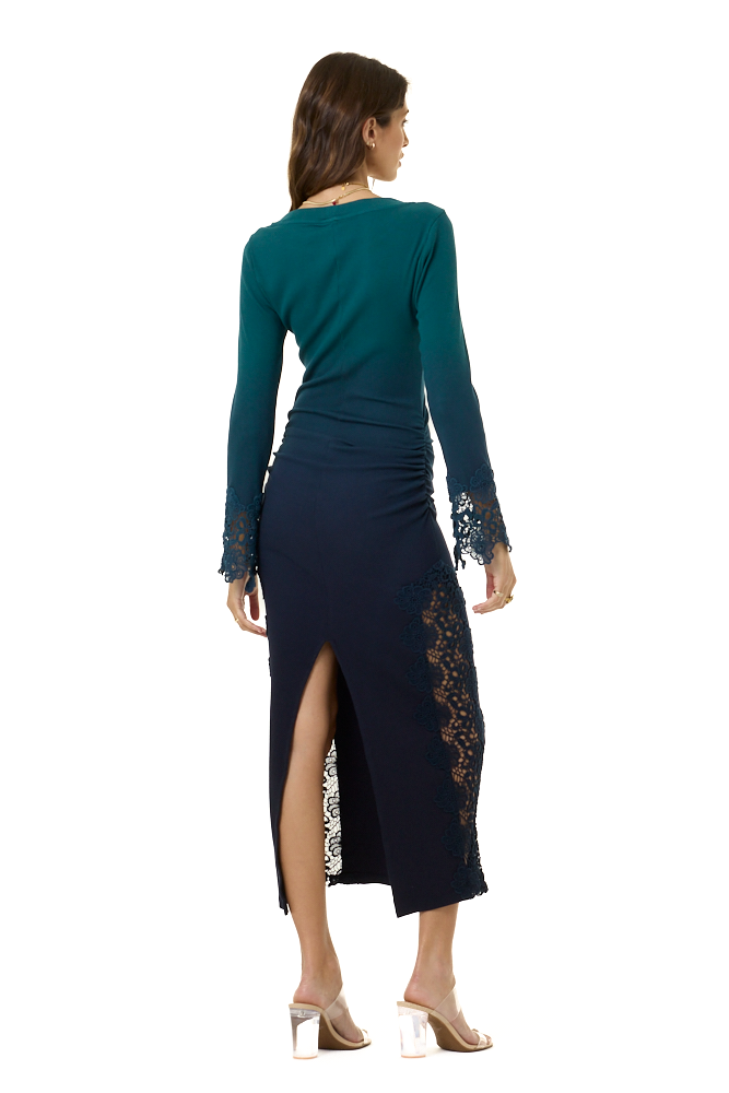 A woman shown from the back wearing a long-sleeve dark teal top and navy midi skirt with lace trim and a side slit, paired with clear high-heeled sandals, against a plain white background.