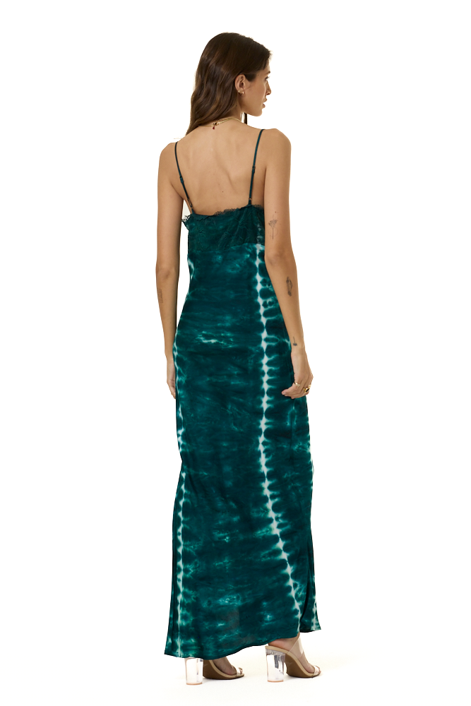 A woman viewed from the back wearing a sleeveless maxi dress with thin straps and a tie-dye pattern in deep teal and white, standing against a plain white background.