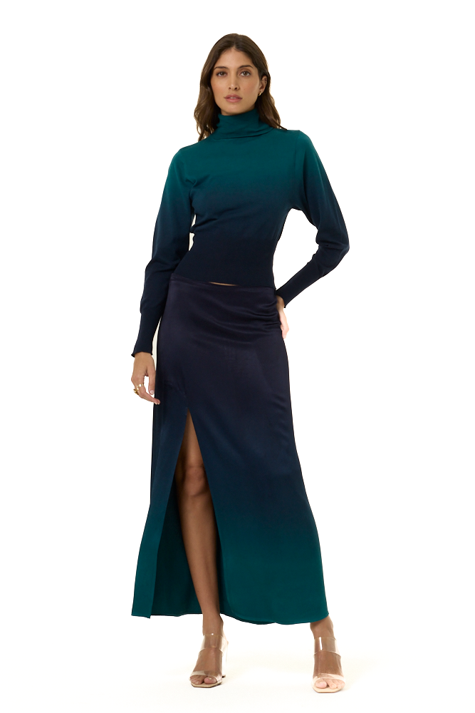 A woman modeling a long-sleeve, high-neck maxi skirt and matching turtle neck sweater with an ombre design in dark teal green and navy blue, featuring a thigh-high slit on the left side, set against a plain white background.