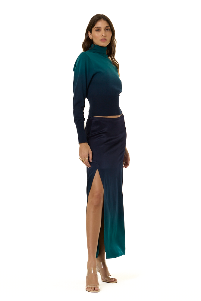 A woman modeling a long-sleeve turtleneck top with a dark teal-to-navy ombre gradient, paired with a matching dark navy to teal ombre skirt, against a plain white background.