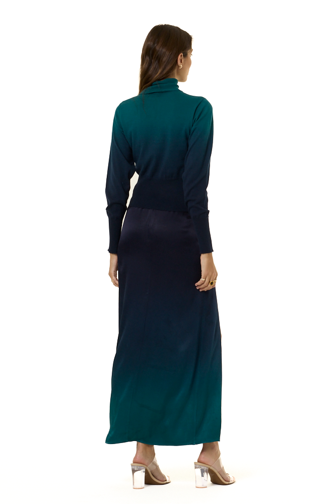A woman from the backside modeling a long-sleeve, high-neck maxi skirt and matching turtle neck sweater with an ombre design in dark teal green and navy blue, featuring a thigh-high slit on the left side, set against a plain white background.
