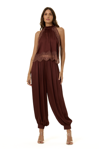 A woman modeling a sleeveless high-neck outfit in deep burgundy with lace trim at the bust and gathered harem-style legs, photographed against a plain white studio background.