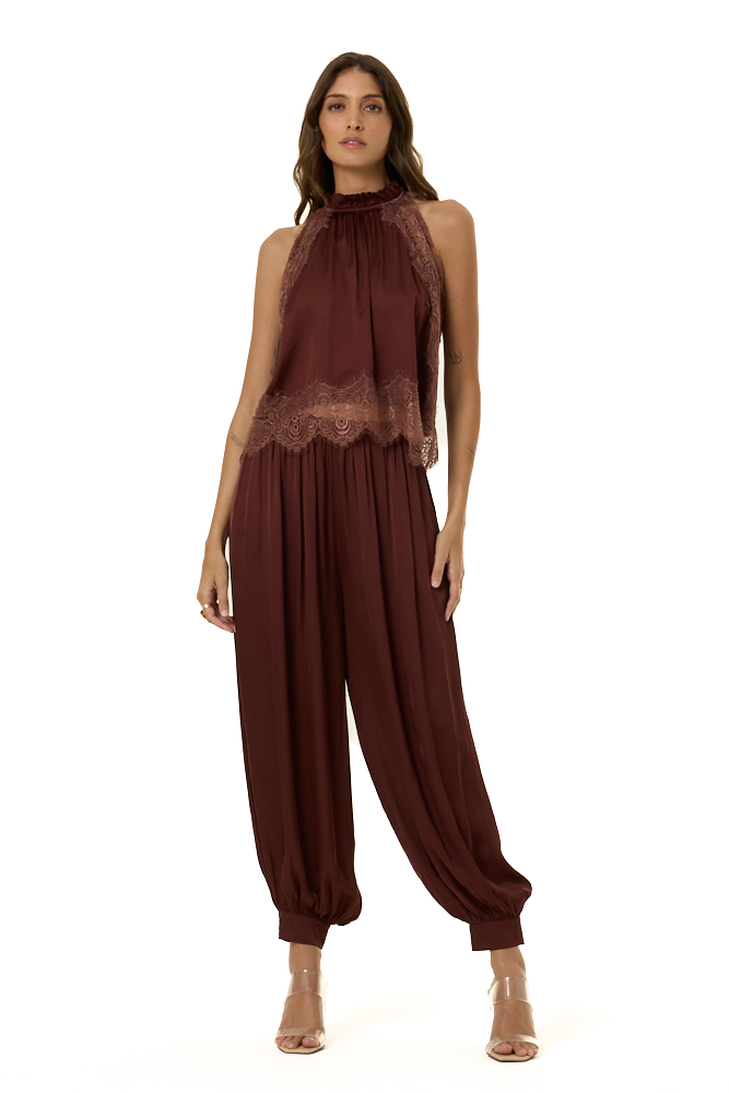 A woman modeling a sleeveless high-neck outfit in deep burgundy with lace trim at the bust and gathered harem-style legs, photographed against a plain white studio background.
