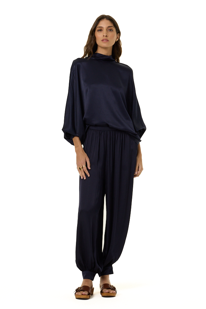 A woman modeling a dark navy blue two-piece outfit consisting of a loose, high-neck long-sleeve top and matching wide-leg pants, paired with brown sandals, against a plain white background.