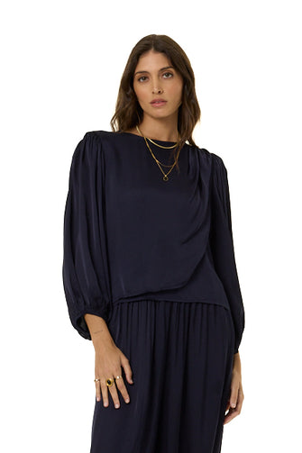 A woman modeling a dark navy blue draped top with voluminous elbow-length sleeves and a matching harem pants styled with gold necklaces and rings against a plain white background.