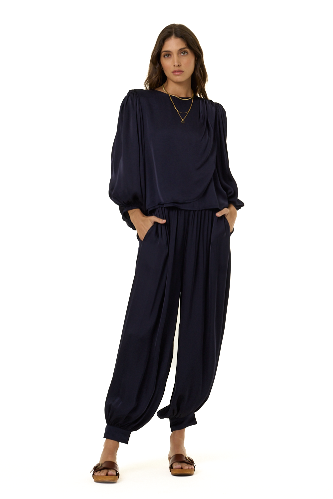 A woman modeling a dark navy blue draped top with voluminous elbow-length sleeves and a matching harem pants styled with gold necklaces and rings against a plain white background.