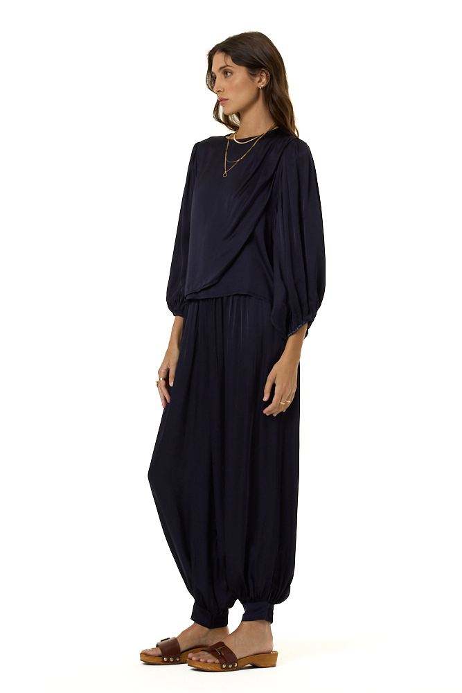 A woman modeling a dark navy blue draped top with voluminous elbow-length sleeves and a matching harem pants styled with gold necklaces and rings against a plain white background.