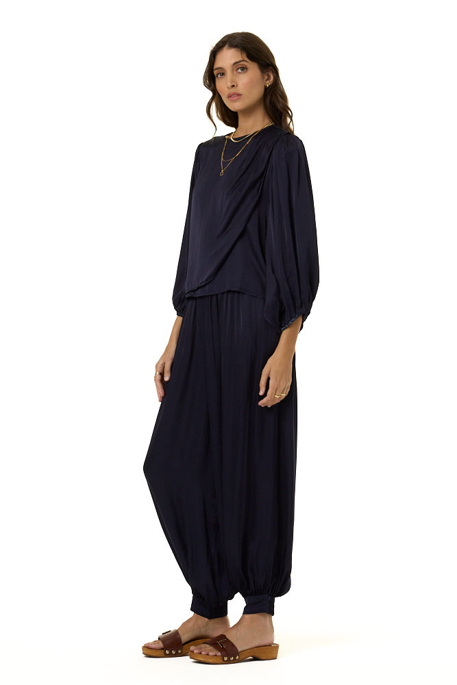 A woman modeling a dark navy blue draped two-piece outfit with voluminous balloon-style pants and long draped sleeves, paired with brown slide sandals, against a plain white background.