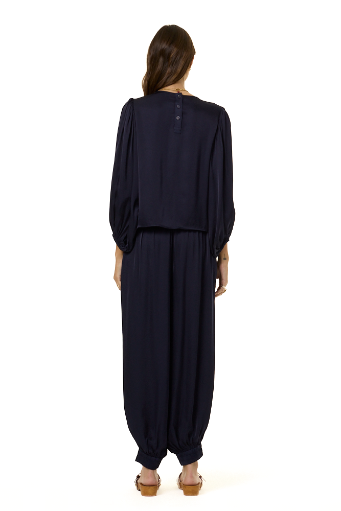 A woman viewed from the back wearing a dark navy blue two-piece outfit with a long-sleeve cropped top and matching loose harem-style pants, paired with tan sandals against a plain white background.