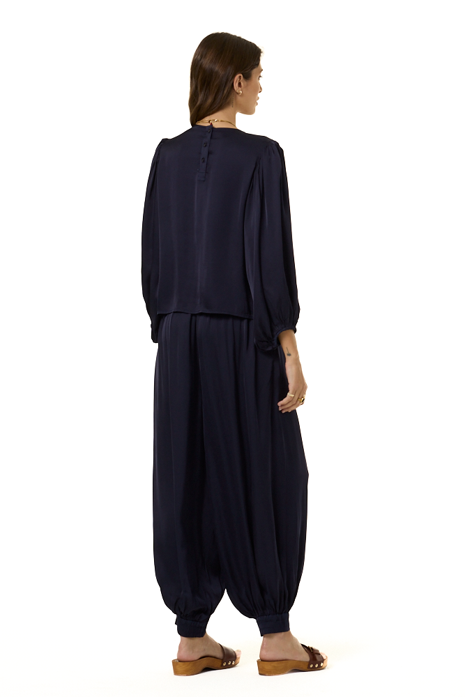 A woman shown from the back wearing a dark navy blue two-piece outfit with a loose long-sleeve top and voluminous harem-style pants, paired with brown sandals, against a plain white background.
