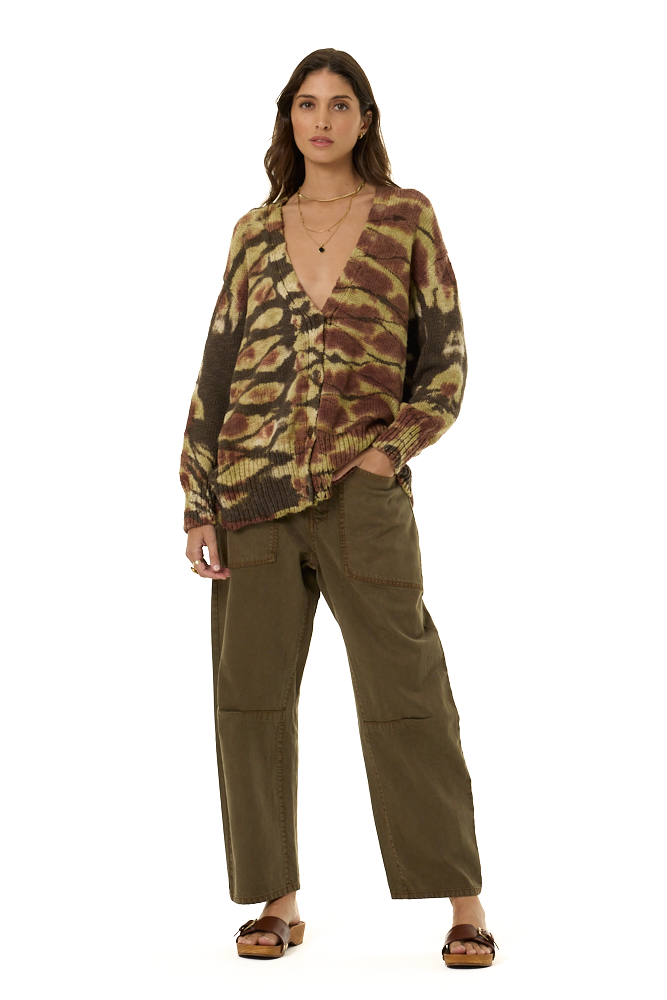 A model stands against a white studio background wearing a brown and olive tie-dye cardigan, olive green wide-leg cargo pants, and brown leather slide sandals.