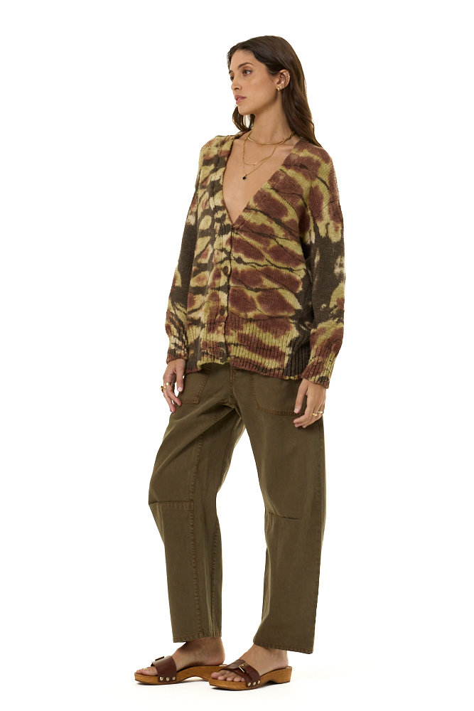 A woman modeling a brown and beige tie-dye knit cardigan sweater with a button-up front, paired with olive green pants, against a solid white background.