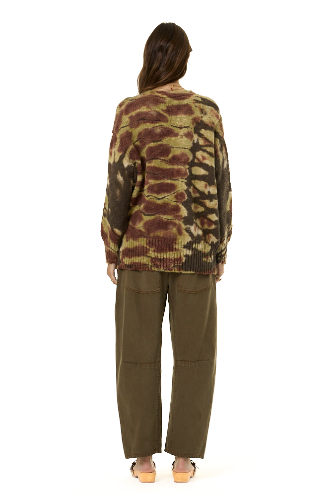 A woman viewed from the back wearing a long-sleeve knit sweater with a brown and olive green abstract tie-dye pattern, paired with olive green wide-leg pants and tan sandals, against a plain white background.