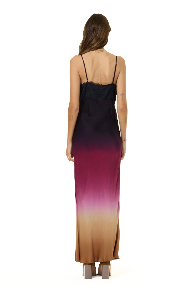A woman viewed from the back wearing a sleeveless maxi dress with thin straps and a gradient color-blocked design transitioning from black at the top to deep magenta and tan at the bottom, set against a plain white background.