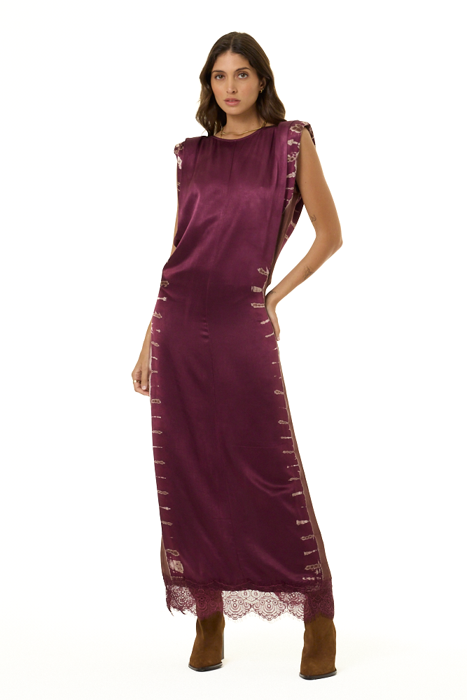 A woman modeling a sleeveless, floor-length plum purple dress with decorative lace trim and side buttons, paired with brown boots, against a plain white background.