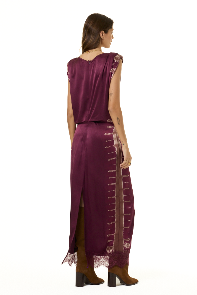 A woman shown from the back wearing a long, sleeveless plum-purple satin dress with lace trim and lace-up detailing down the side, paired with brown boots, against a plain white background.
