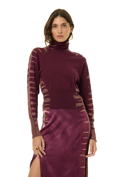 A woman modeling a long-sleeve turtleneck dress in deep plum purple, featuring textured knit sleeves and a satin-like skirt, set against a plain white background.